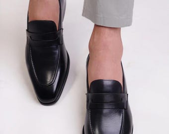 Op de afbeelding: Zwarte leren loafers met een klassiek design. De schoenen hebben een gladde, glanzende afwerking en een instapmodel met een banddetail over de bovenkant. De loafers worden vanuit een lage hoek getoond, waardoor hun slanke vorm wordt benadrukt.