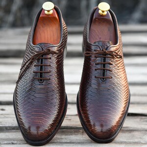Könnte beinhalten: Ein Paar braune Lederschuhe mit strukturiertem Muster und braunen Schnürsenkeln. Die Schuhe werden mit hölzernen Schuhspannern präsentiert. Sie haben ein klassisches, formelles Design.