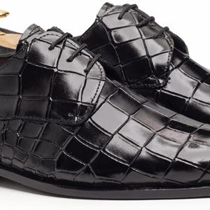 May include: A pair of black dress shoes with a glossy crocodile-embossed texture, featuring black laces, a rounded toe, and a low heel. Each shoe contains a wooden shoe tree with a gold-toned metal knob handle inserted into the heel area.