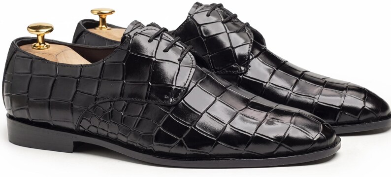 May include: A pair of black dress shoes with a glossy crocodile-embossed texture, featuring black laces, a rounded toe, and a low heel. Each shoe contains a wooden shoe tree with a gold-toned metal knob handle inserted into the heel area.