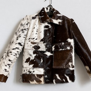 May include: A patchwork jacket with a cowhide design. The jacket features a mix of brown and white spotted patterns, with two front pockets and button closures. The jacket is hanging on a black hanger.