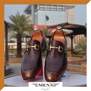 May include: A pair of dark brown loafers with a snakeskin-like texture and a gold-toned metal horsebit detail across the vamp. Each shoe features a wooden shoe tree insert inside the opening. The text "MEN'S Carlo" is printed at the bottom.
