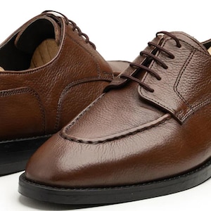 May include: A pair of brown leather dress shoes with dark brown laces and black soles. The shoes have a classic design with a textured leather upper and are shown with wooden shoe trees.