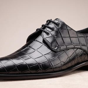 May include: A black dress shoe with a glossy finish and a textured crocodile skin pattern. The shoe features a rounded toe, a low heel, and black laces threaded through four eyelets on each side of the central tongue area.