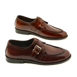 May include: Two pairs of brown leather dress shoes with a glossy finish. Each shoe features a strap with a gold-colored buckle and decorative perforations along the edges. The shoes have a classic, formal design.