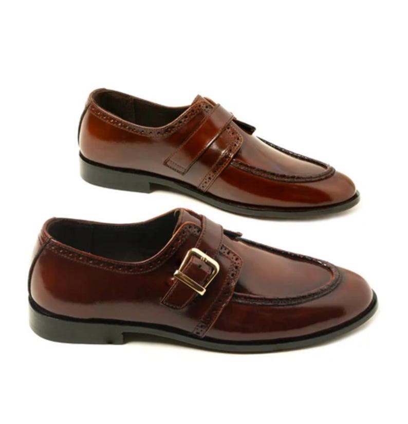 May include: Two pairs of brown leather dress shoes with a glossy finish. Each shoe features a strap with a gold-colored buckle and decorative perforations along the edges. The shoes have a classic, formal design.
