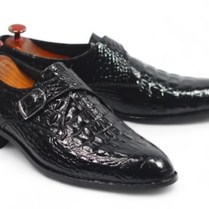 K&ouml;nnte beinhalten: Ein Paar schwarze Lederschuhe mit Krokodilleder-Textur. Die Schuhe haben einen Schnallenverschluss und einen niedrigen Absatz. Schuhspanner sind in die Schuhe eingelegt. Auf wei&szlig;em Hintergrund.