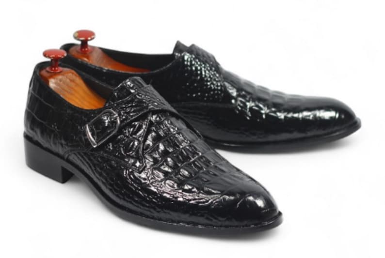 K&ouml;nnte beinhalten: Ein Paar schwarze Lederschuhe mit Krokodilleder-Textur. Die Schuhe haben einen Schnallenverschluss und einen niedrigen Absatz. Schuhspanner sind in die Schuhe eingelegt. Auf wei&szlig;em Hintergrund.