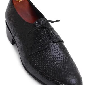 Può includere: Scarpa da uomo in pelle nera con motivo strutturato simile a quello di un rettile. La scarpa ha una chiusura con lacci e un piccolo tacco. All'interno è presente una forma per scarpe in legno con pomello dorato.