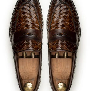 May include: A pair of dark brown leather loafers featuring a woven texture across the upper and a solid leather strap across the vamp. Each shoe contains a light-coloured wooden shoe tree with a metallic knob at the heel for maintaining shape.