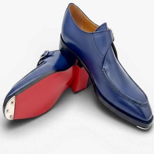 May include: A pair of blue leather monk strap shoes featuring a smooth finish, a side buckle closure, and a vibrant red sole. The front of the sole is reinforced with a silver-toned metal toe plate, and the interior lining is a light tan color.