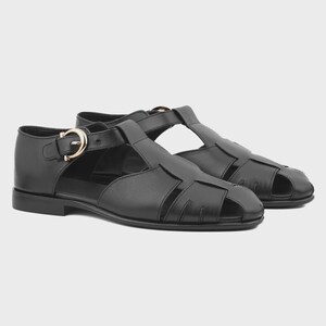 May include: A pair of black leather fisherman sandals featuring a closed-toe design with wide woven straps, a T-strap front, and an adjustable ankle strap secured by a polished gold-tone metal buckle. The sandals have a low, flat heel.