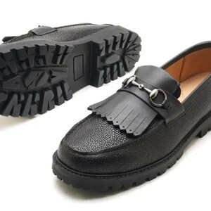 May include: A pair of black loafers with a pebbled texture, featuring a fringed kiltie detail across the vamp and a silver-toned metal horsebit ornament. The shoes have a thick, lugged rubber sole with a deep tread pattern for traction.