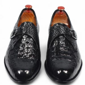 Könnte beinhalten: Ein Paar schwarze Lederschuhe mit Monk-Strap-Verschluss und Krokodilhautstruktur. Die Schuhe haben ein glänzendes Finish und einen Schnallenverschluss. Holzschuhspanner mit roten Oberteilen befinden sich im Inneren.