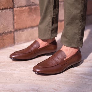 May include: A pair of brown leather penny loafers featuring a textured vamp and a smooth heel counter. The shoes have a rounded toe, a low heel, and a classic slip-on design. The leather has a polished finish, and the shoes are worn with olive green trousers.