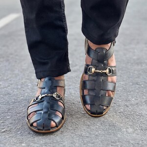 May include: Black leather fisherman sandals featuring a textured grain finish, a closed-toe cage design, and a gold-toned metal horsebit detail across the instep. The sandals include an adjustable ankle strap with a buckle and a flat brown sole.