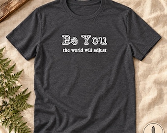 May include: A dark charcoal gray short-sleeve t-shirt featuring the white text "Be You" in a bold, outlined font, followed by the smaller white text "the world will adjust" centered on the chest area of the garment.
