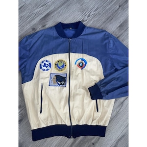 Peut inclure: Veste zippée vintage bleue et crème avec des écussons BMW. La veste a un col, des poignets et des manches supérieures bleus, avec un panneau avant crème. Les écussons incluent des logos et du texte BMW. Deux poches avant.