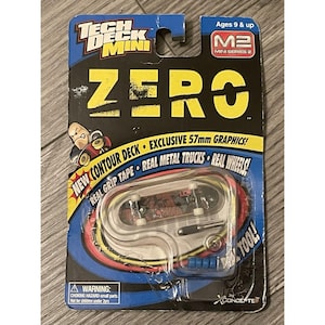 Pode incluir: Um brinquedo de skate Tech Deck Mini, embalado em um cartão azul e amarelo. A embalagem apresenta a palavra "ZERO" em letras amarelas grandes e inclui um skate em miniatura com um design gráfico, trucks de metal reais e rodas reais.