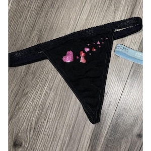 May include: Black thong underwear with a decorative heart design in pink glitter. The underwear has a black lace trim and a light blue elastic band with the word "SECRET" printed on it.
