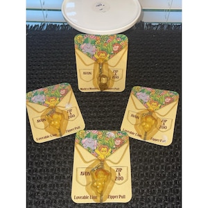 Op de afbeelding: Vier Avon "Zip A Zoo" ritstrekkers. Elke trekker heeft een kleine, goudkleurige dierenbedel bevestigd aan een kaart met een kleurrijk jungle-ontwerp. De kaarten lezen "Loveable Lion" en "Modest Monkey".