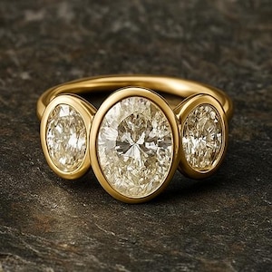 Könnte beinhalten: Ein goldener Ring mit drei oval geschliffenen Diamanten. Der mittlere Diamant ist größer als die beiden seitlichen Steine. Der Ring liegt auf einer dunklen, strukturierten Oberfläche, die das Funkeln der Edelsteine hervorhebt.
