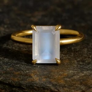 May include: A gold ring featuring a rectangular, pale blue gemstone. The gemstone is set with four prongs, and the band is a simple, polished gold. The ring is displayed on a dark, textured surface.