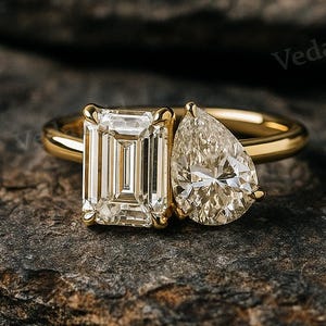 May include: A gold ring with two diamond stones. One is an emerald cut, the other a pear-shaped diamond. The ring is set against a dark, textured background.