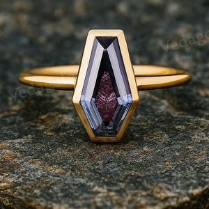 Pode incluir: Um anel de ouro com uma gema de formato geométrico único, com tons roxos e azuis. O anel tem uma banda de ouro fina, e a gema é engastada em uma moldura de ouro. O anel é exibido em uma superfície cinza texturizada.