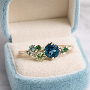 Può includere: Un anello d'oro con un gruppo di pietre preziose blu, verdi e bianche, presentato in un portagioie di velluto blu chiaro. L'anello presenta una grande gemma rotonda blu scuro come punto focale.