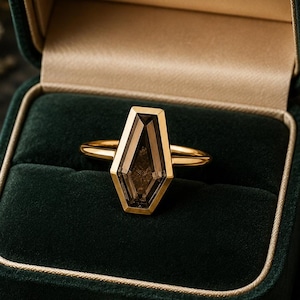 May include: A gold ring with a geometric, smoky-colored gemstone is displayed in a dark green velvet ring box. The ring's design features a unique, elongated shape. The box is open, revealing a gold interior and a small, gold clasp.