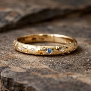 Puede incluir: Un anillo de oro con un diseño celestial. El anillo presenta estrellas grabadas y está adornado con pequeñas piedras preciosas de varios colores, incluyendo blanco, amarillo y azul. El anillo está sobre un fondo texturizado y oscuro.