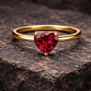 May include: A gold ring with a heart-shaped, deep red gemstone. The ring is set on a textured, dark gray stone surface. The gemstone is faceted, catching the light and creating a sparkling effect.