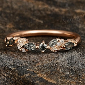 May include: Rose gold ring with a delicate design. The ring features marquise-cut and round green gemstones, along with small white gemstones. The ring is set on a textured surface.