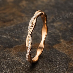 Op de afbeelding: Een roségouden ring met een gedraaid bandontwerp. De ene helft van de band is glad en gepolijst, terwijl de andere helft is bezet met een rij kleine, heldere, rondgeslepen stenen. De ring staat rechtop en toont zijn verweven structuur.