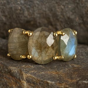May include: A gold ring featuring three oval-cut labradorite gemstones. The gemstones are a mix of gray and blue hues, set in gold prongs. The ring is displayed on a textured, dark gray surface.