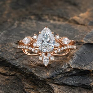 May include: A rose gold ring set featuring a large pear-cut center stone surrounded by smaller round and pear-shaped accent stones. The band has a curved, layered design with pavé-set diamonds, creating a multi-stone cluster appearance.
