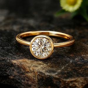 May include: A gold ring with a large, round, clear gemstone set in a bezel. The ring is on a dark, textured surface. The gemstone sparkles, reflecting light. The ring's band is smooth and polished.