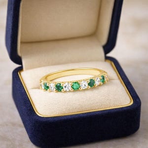 Puede incluir: Un anillo de banda de oro con piedras preciosas verdes esmeralda y claras alternas. El anillo se muestra dentro de un joyero de terciopelo azul con un interior color crema. Una delicada pieza de joyería.