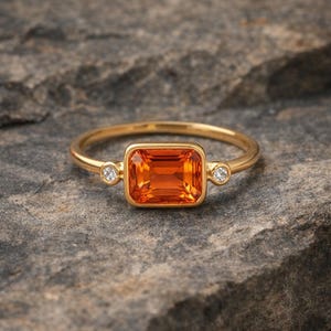 May include: A gold ring with an orange rectangular gemstone as the centerpiece. Two small, round, clear gemstones flank the main stone. The ring is set against a gray, textured background.