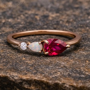 May include: A rose gold ring featuring three gemstones: a small, round diamond, a pear-shaped opal, and a large, pear-shaped ruby. The ring is set against a textured, brown background.