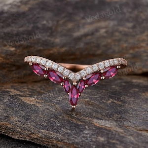 May include: A rose gold ring with a V-shaped design. The ring features a row of small, clear diamonds along the top and a series of marquise-cut, deep red gemstones. The ring is set against a dark, textured background.