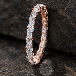 Puede incluir: Anillo de eternidad de oro rosa con una fila continua de piedras preciosas redondas y claras. El anillo se encuentra sobre un fondo oscuro y texturizado, resaltando el brillo de las piedras. El anillo es una joya.