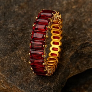 Könnte beinhalten: Ein goldener Ring mit einer durchgehenden Reihe tiefroter, rechteckiger Edelsteine im Baguetteschliff, die in einer Krappenfassung gehalten werden. Der Ring zeigt einen lebendigen Kontrast zwischen dem polierten Gold und den dunkelroten Steinen.