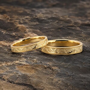May include: Two yellow gold rings featuring an intricate engraved floral scroll pattern along the outer band. Each ring has a polished finish with a beaded edge detail, showcasing a continuous embossed design that wraps around the entire circumference.