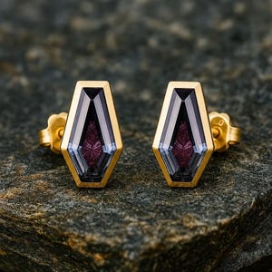 May include: A pair of stud earrings featuring a coffin-shaped design with a gold-toned metal setting. Each earring contains a faceted, translucent purple gemstone at the center, surrounded by a dark, smoky-colored border within the gold frame.
