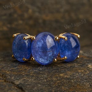 Puede incluir: Un anillo de oro con tres piedras preciosas ovales de color azul intenso. El anillo tiene una banda de oro con delicadas garras. Las piedras preciosas están engastadas en una banda de oro. El anillo se muestra sobre una superficie texturizada marrón oscuro.