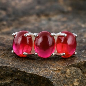 以下が含まれることがあります： 3つの楕円形の鮮やかな赤い宝石をあしらったシルバーリング。宝石は一列に並んでおり、磨かれたシルバーバンドと各石を固定する爪が付いています。リングは、質感のある灰色の表面に表示されています。