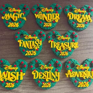 May include: Eight green wreath ornaments shaped like Mickey Mouse ears, each featuring red berries and yellow text. The ornaments display the word Disney, a unique name like Magic, Wonder, Dream, Fantasy, Treasure, Wish, Destiny, or Adventure, and 2026.