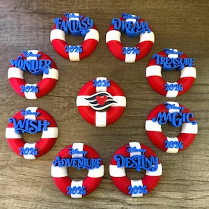 May include: A collection of red, white, and blue life preserver-shaped ornaments. Each ornament features a different Disney cruise ship name in blue, such as "Fantasy" and "Dream", with the year "2026" or "2020".